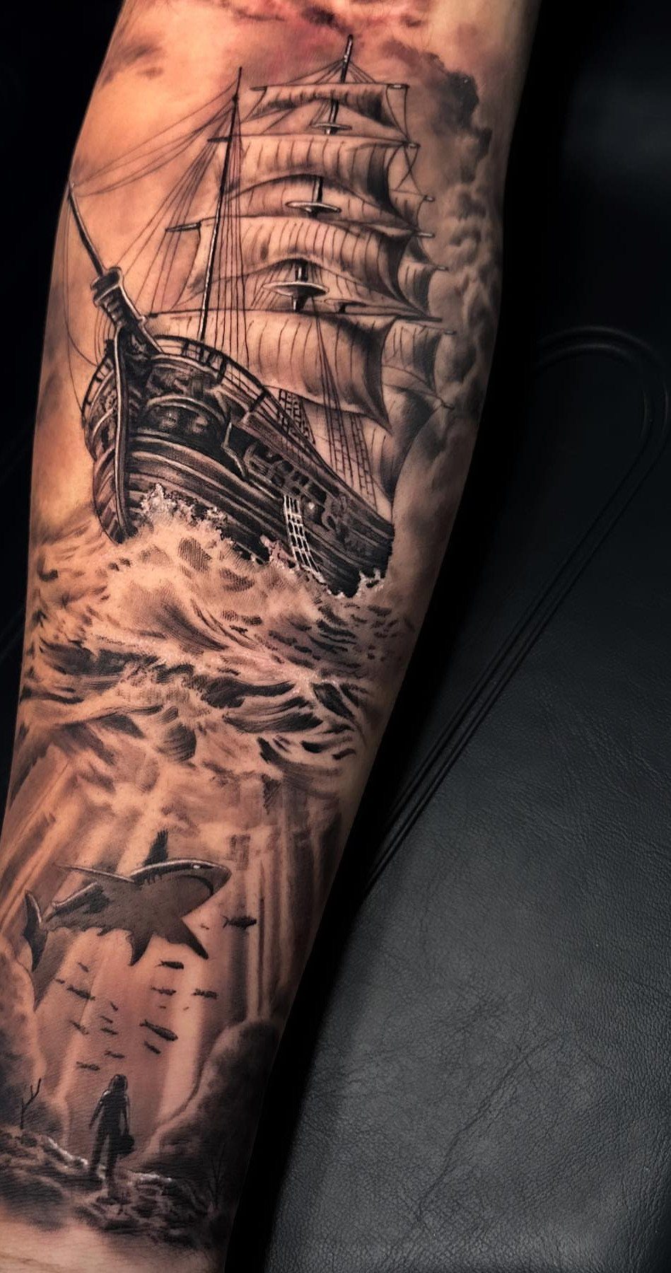 16 Ship Tattoo Ideas in Anchor, Viking, Pirate and Seascape Styles 8 A detailed black and grey realistic ship tattoo on a person's arm with waves and a shark.