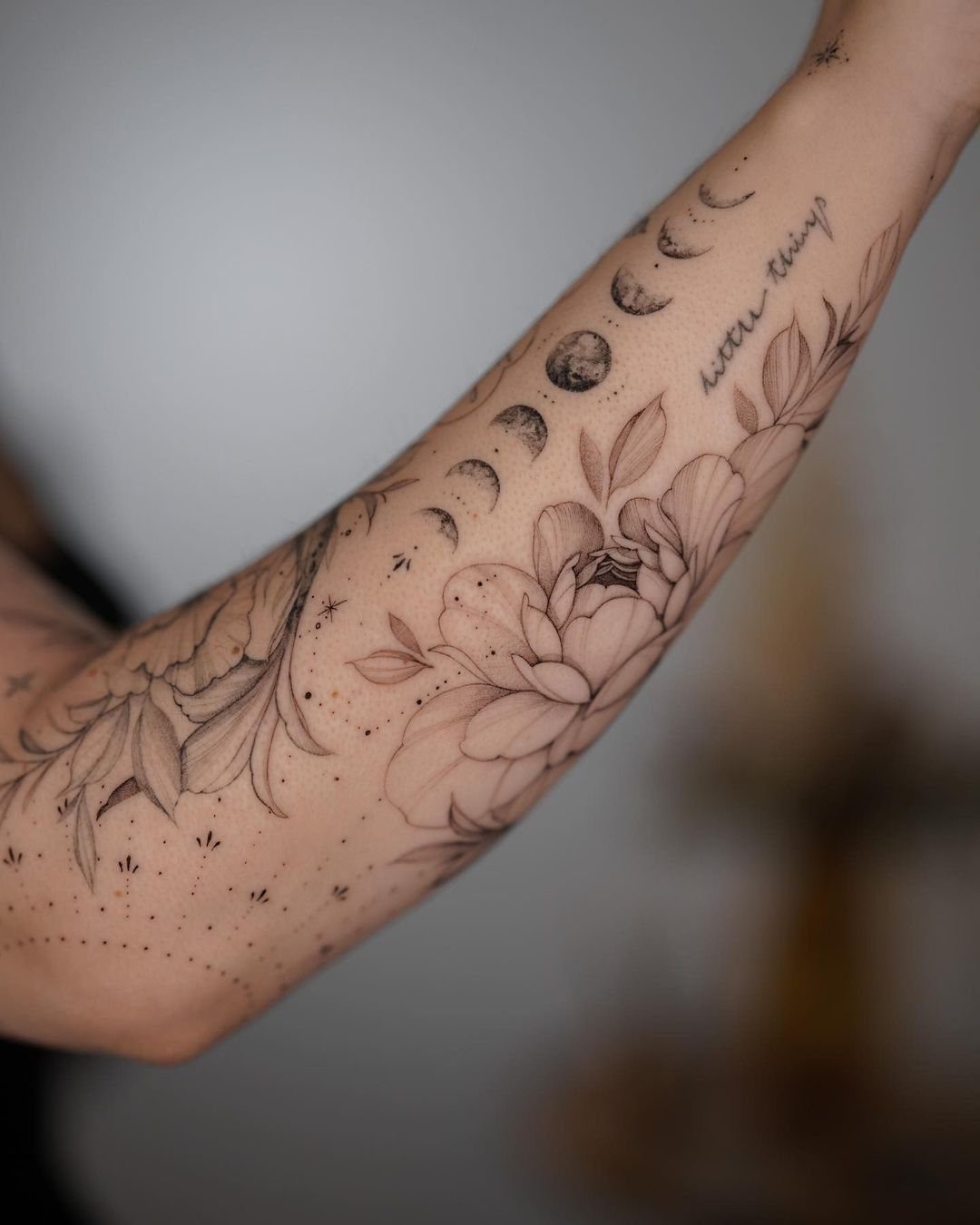 7 Simple Moon Phase Tattoo Designs for Every Style 4 A forearm tattoo showing moon phases in an arc above large botanical flowers and leaves.