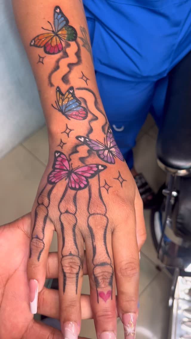 7 Skeleton Hand Tattoo Ideas for Your Next Ink 5 A hand tattoo featuring black line-art skeleton bones and several colorful butterflies on the skin.
