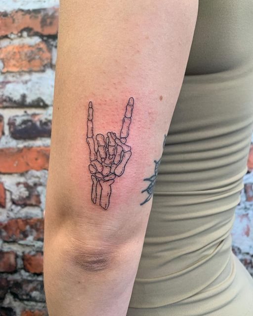 7 Skeleton Hand Tattoo Ideas for Your Next Ink 2 A fine-line black ink tattoo of a skeleton hand making the horns gesture on an arm.