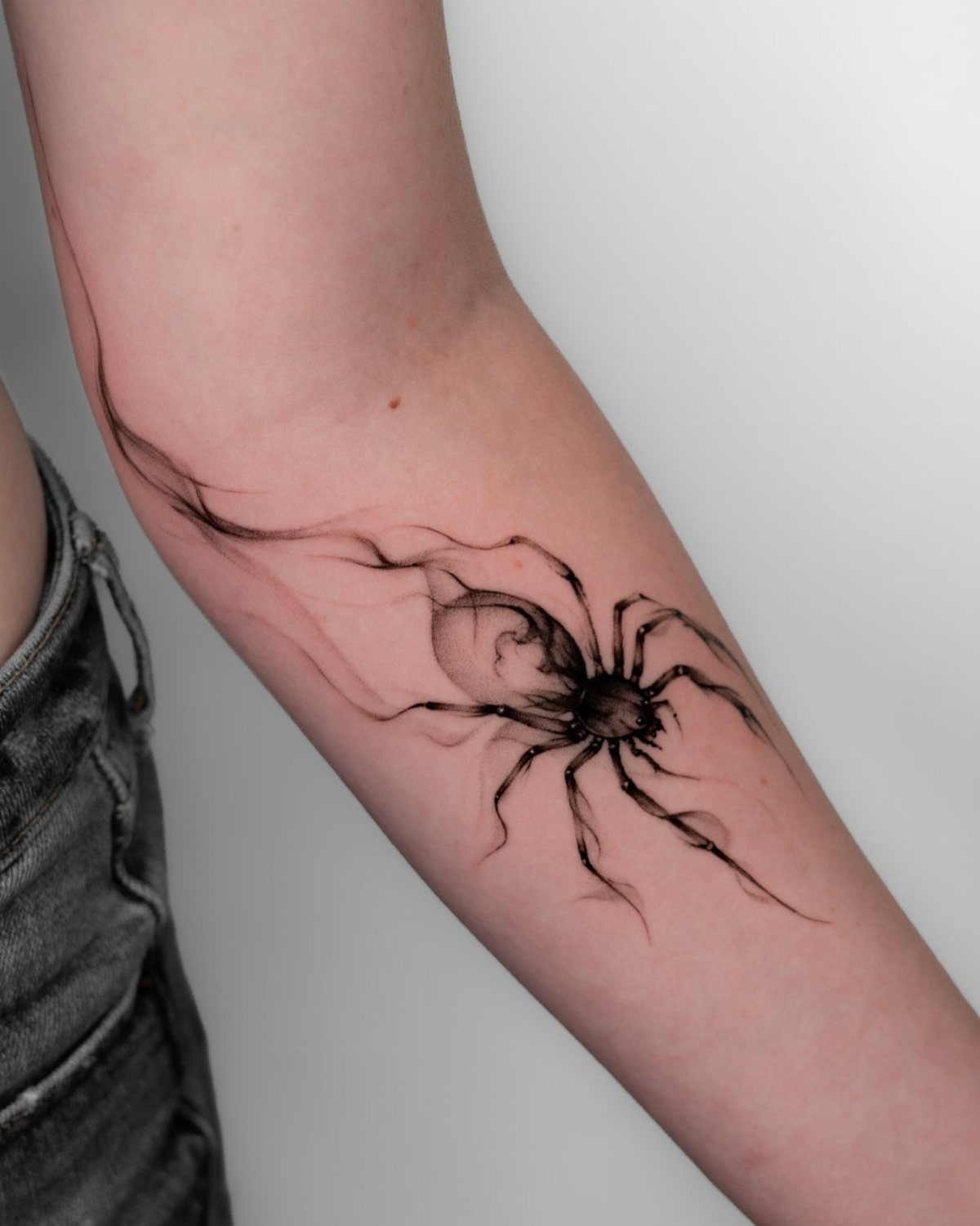 12 Spider Tattoo Ideas That Balance Creepy and Beautiful 10 A large black spider tattoo with wispy, smoke-like legs on a person's forearm.