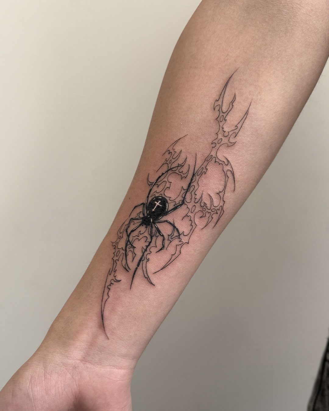 12 Spider Tattoo Ideas That Balance Creepy and Beautiful 11 A black ink spider tattoo with a cross on its back on a person's forearm.