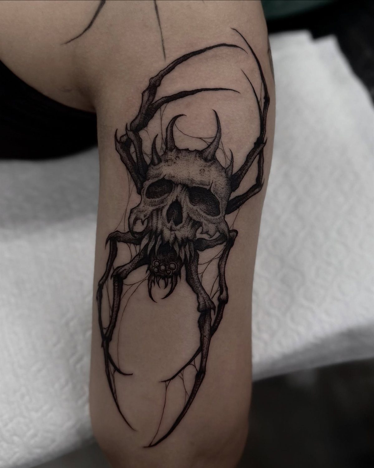 12 Spider Tattoo Ideas That Balance Creepy and Beautiful 6 A black ink tattoo of a spider with a decayed skull face on a person's arm.