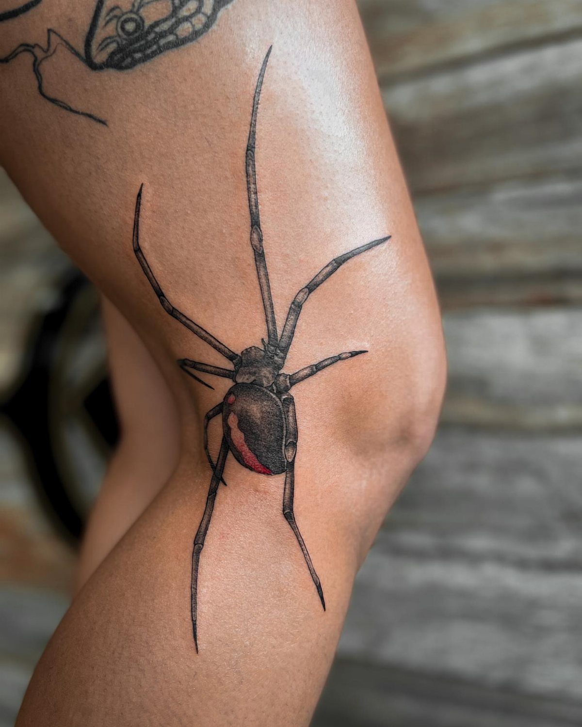 12 Spider Tattoo Ideas That Balance Creepy and Beautiful 3 A black and red spider tattoo inked on a person's leg near the knee.