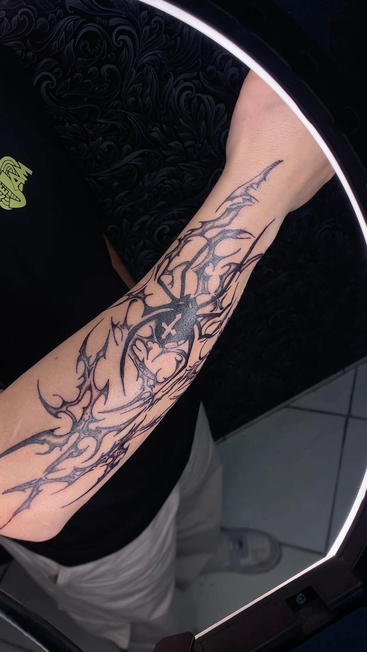 12 Spider Tattoo Ideas That Balance Creepy and Beautiful 1 A black ink tribal spider tattoo with sharp, flowing patterns on a person's forearm.