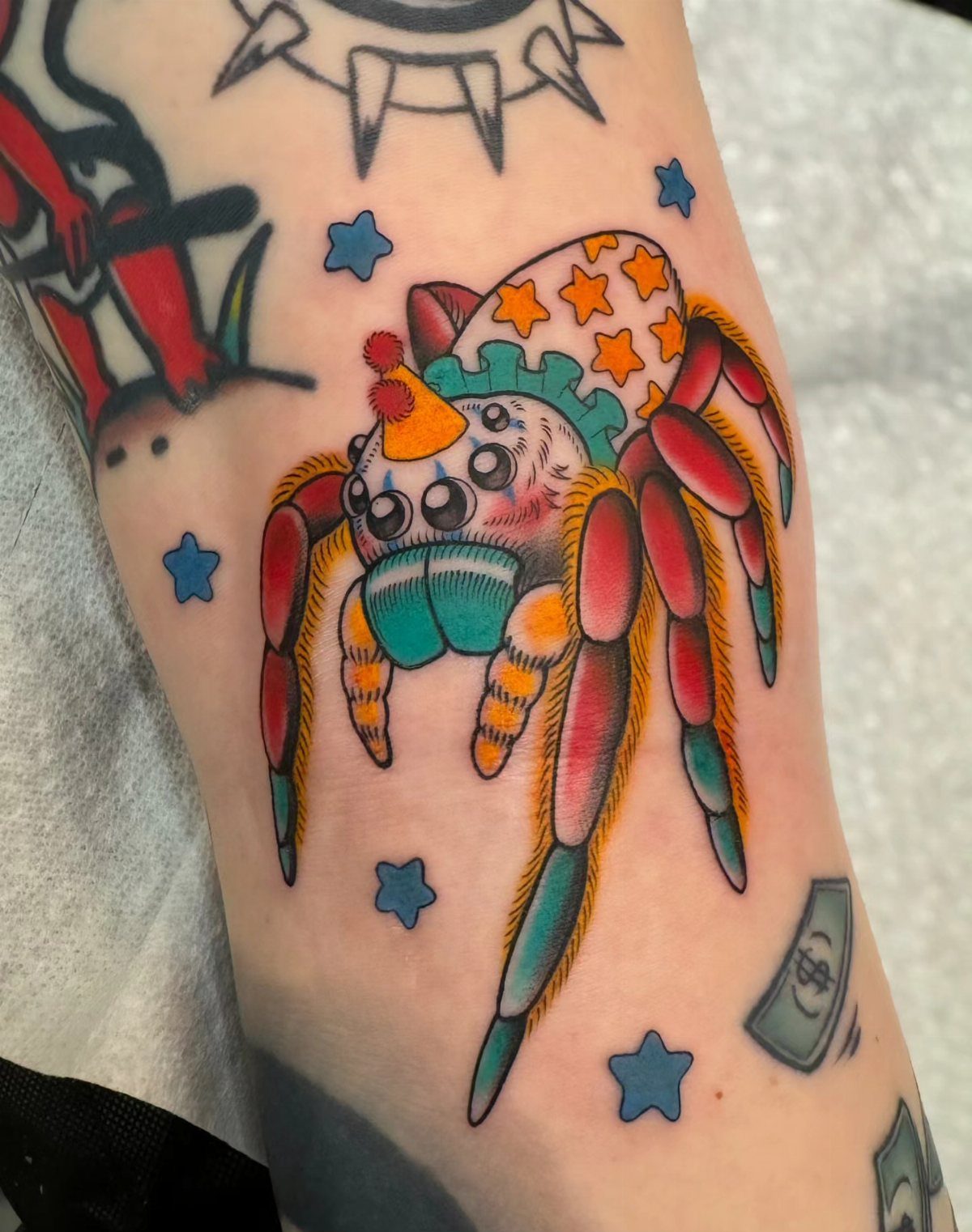 12 Spider Tattoo Ideas That Balance Creepy and Beautiful 7 A colorful illustrative spider tattoo with stars and a tiny party hat on skin.
