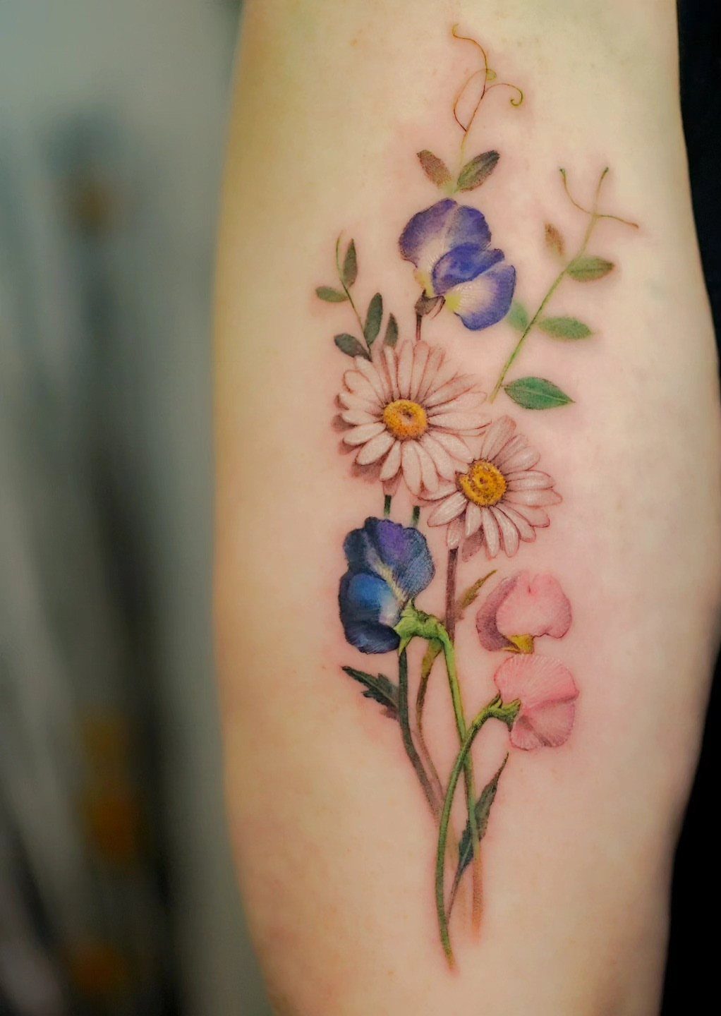 Sweet Pea Flower Tattoo: 7 Ideas for Placement and Style 4 Watercolor tattoo of blue sweet pea flowers and white daisies on a person's skin.