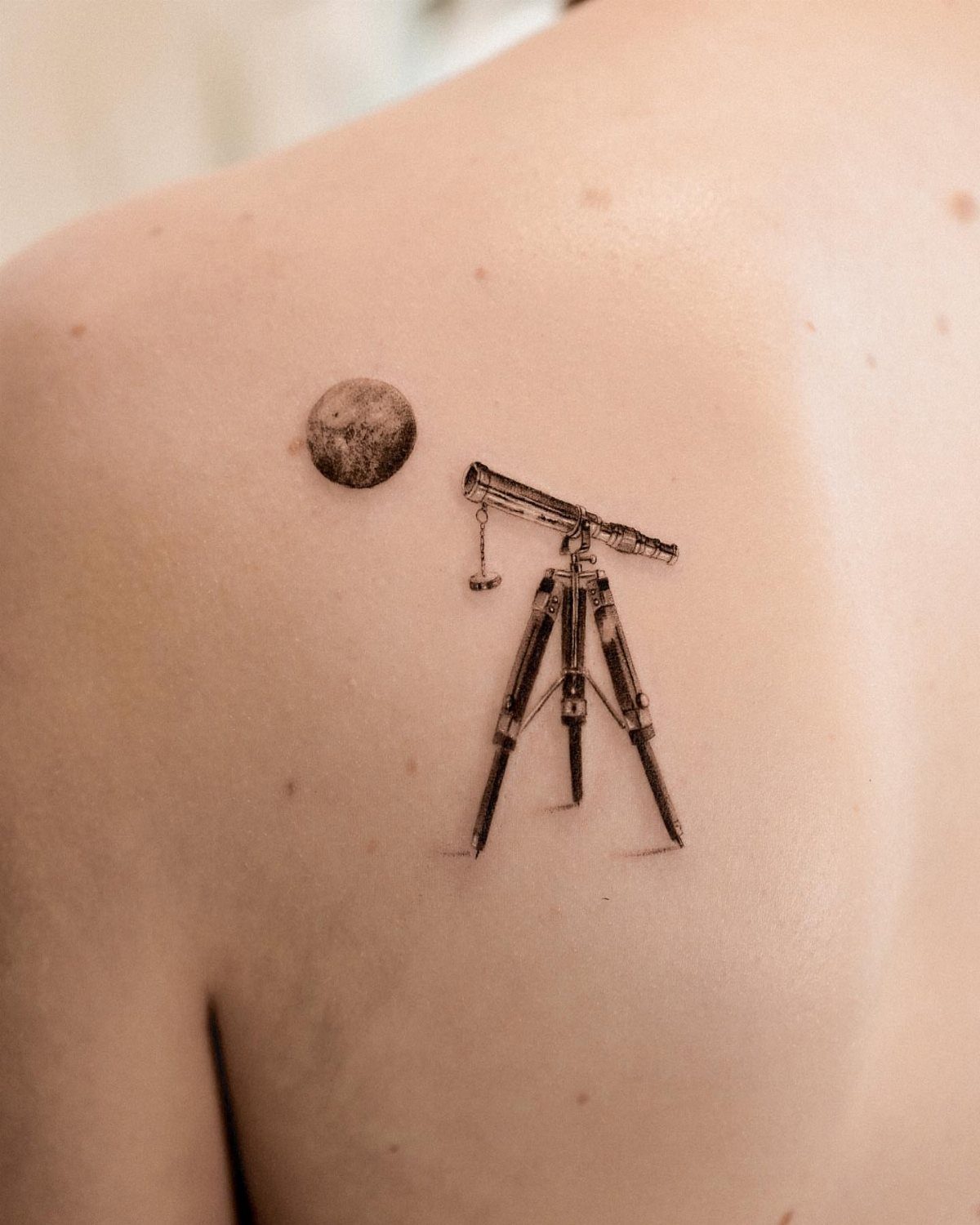 7 Telescope Tattoo Ideas for Every Space and Style 1 A small black ink tattoo of a telescope on a tripod and a moon on a shoulder.