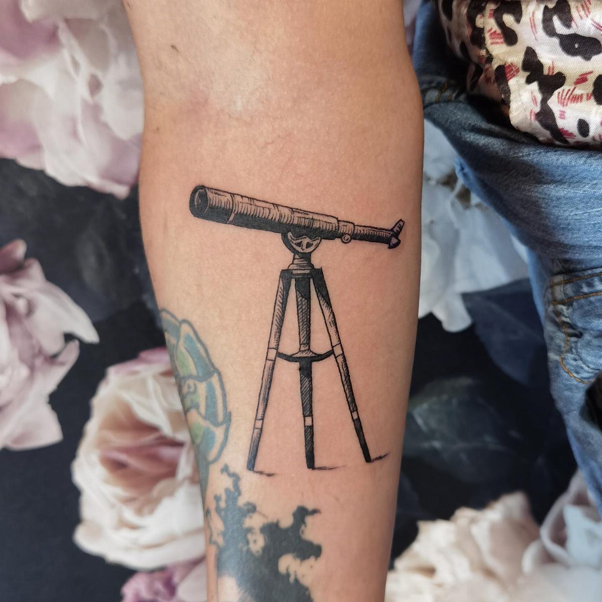7 Telescope Tattoo Ideas for Every Space and Style 5 A black ink tattoo of a vintage telescope on a tripod on a person's arm.