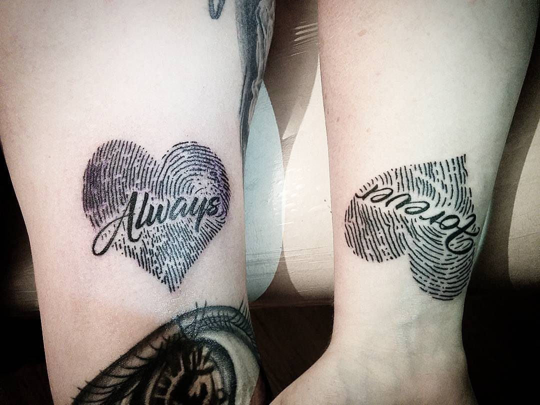 6 Thumbprint Heart Tattoo Ideas to Seal a Bond in Ink 2 Two black ink thumbprint heart tattoos with cursive text on skin.