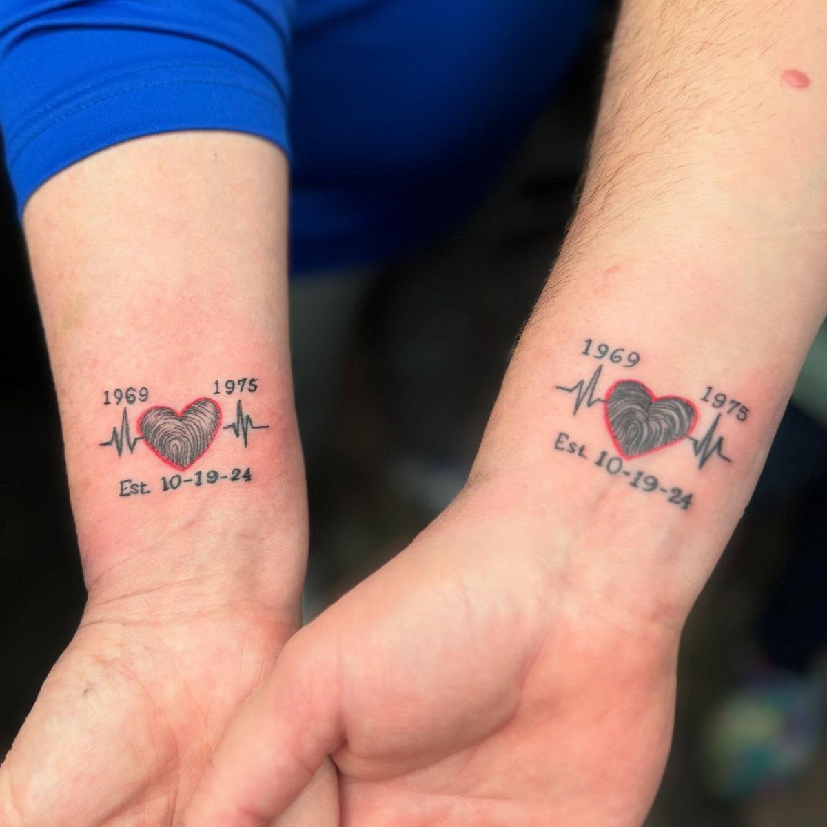 6 Thumbprint Heart Tattoo Ideas to Seal a Bond in Ink 3 Two wrists displaying matching heart tattoos made of fingerprints with red and black ink and dates.