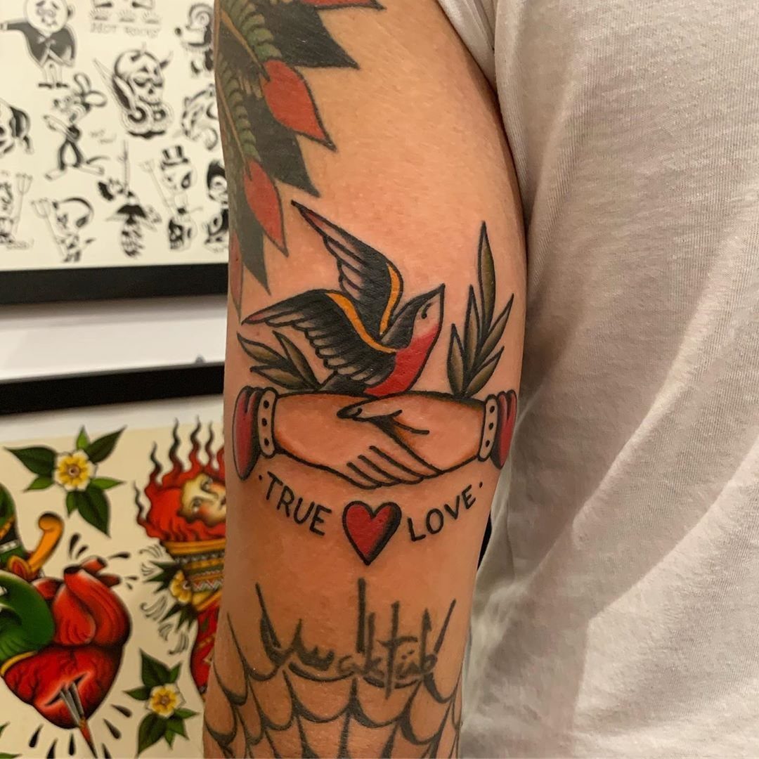 Traditional Handshake Tattoo: 9 Designs From Heart Frames to Skeleton Hands 6 Traditional tattoo on an arm showing hands shaking with a swallow bird and a love banner.