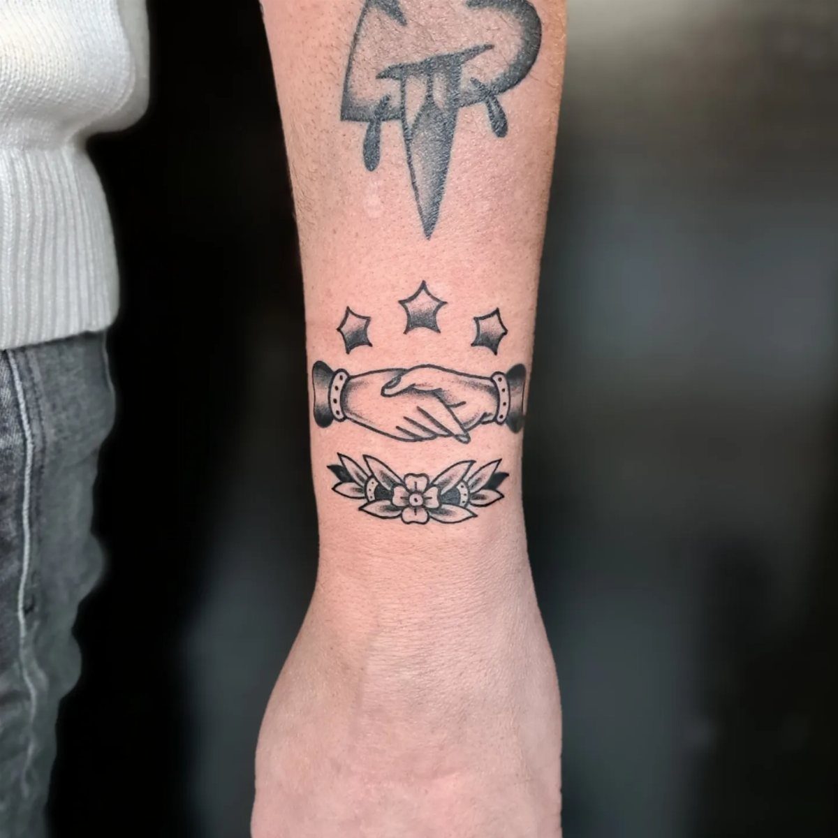 Traditional Handshake Tattoo: 9 Designs From Heart Frames to Skeleton Hands 9 Black ink traditional style tattoo of two hands shaking with stars and flowers on a forearm.