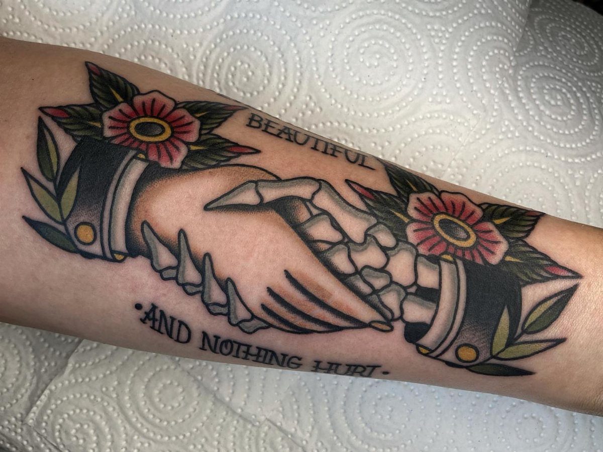 Traditional Handshake Tattoo: 9 Designs From Heart Frames to Skeleton Hands 3 Skeletal hands shaking in an American traditional tattoo style with red flowers and text.