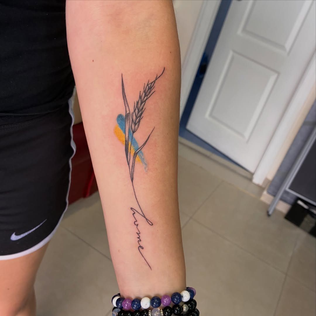 6 Wheat Tattoo Ideas for Your Next Ink 6 A fine line wheat stalk tattoo with blue and yellow watercolor splashes on a forearm.