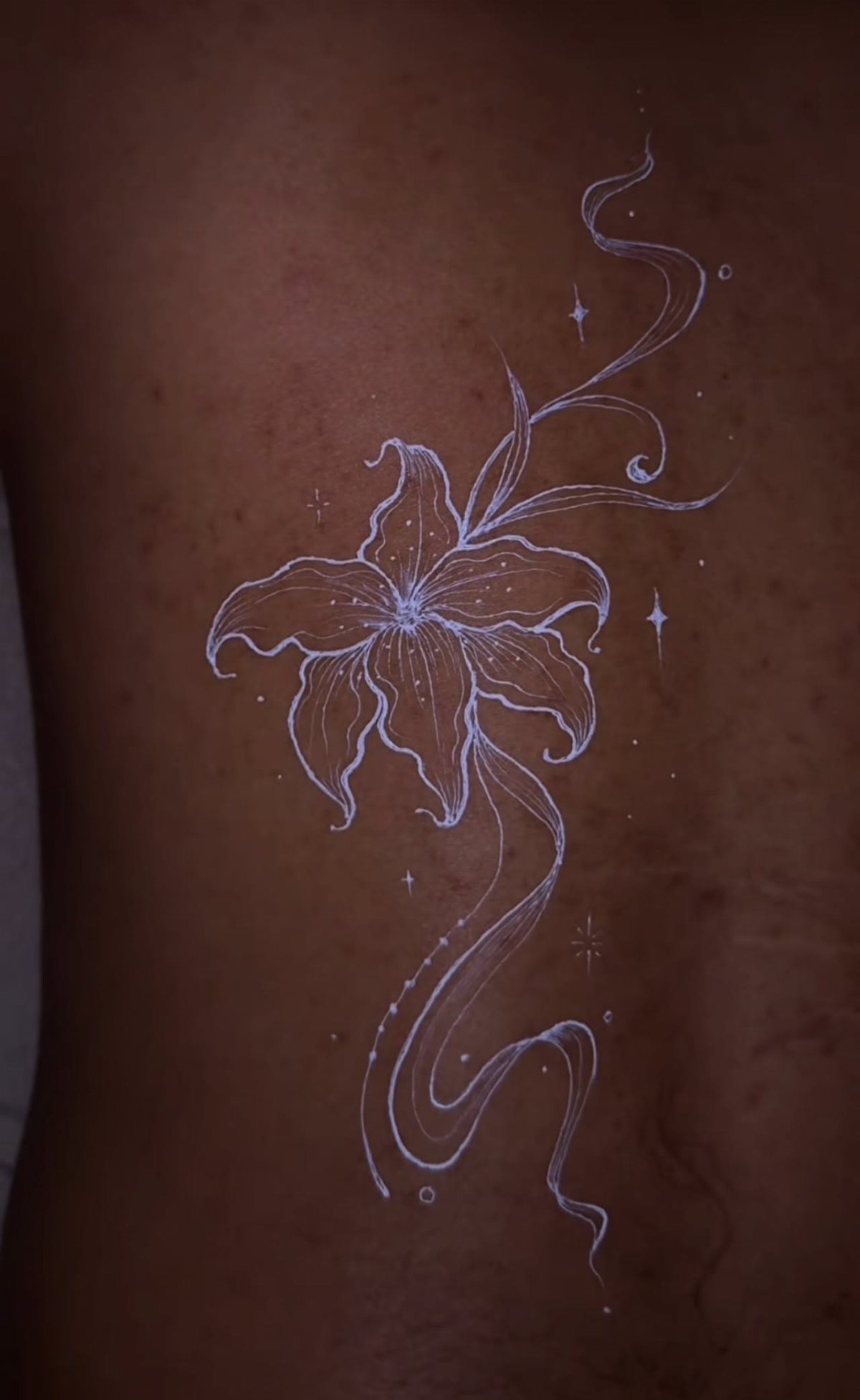White Ink Water Lily Tattoo: 7 Ideas for Subtle Floral Ink 4 White ink water lily tattoo with flowing stems and star accents on dark skin.
